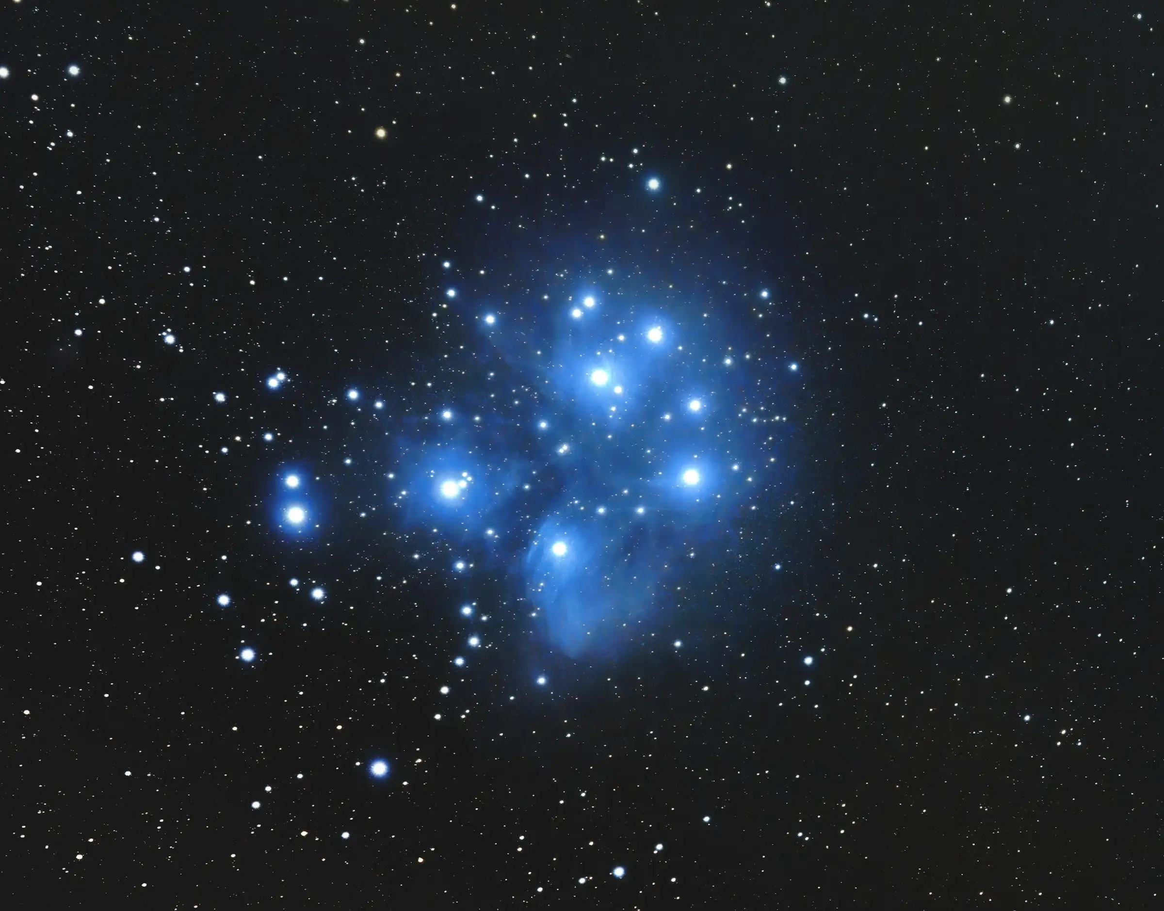 The Pleiades star cluster, a bright group of blue stars wrapped in nebulosity against a black sky.